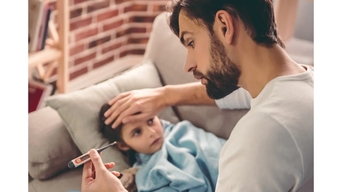 A father takes the temperature of a small child lying on a couch.