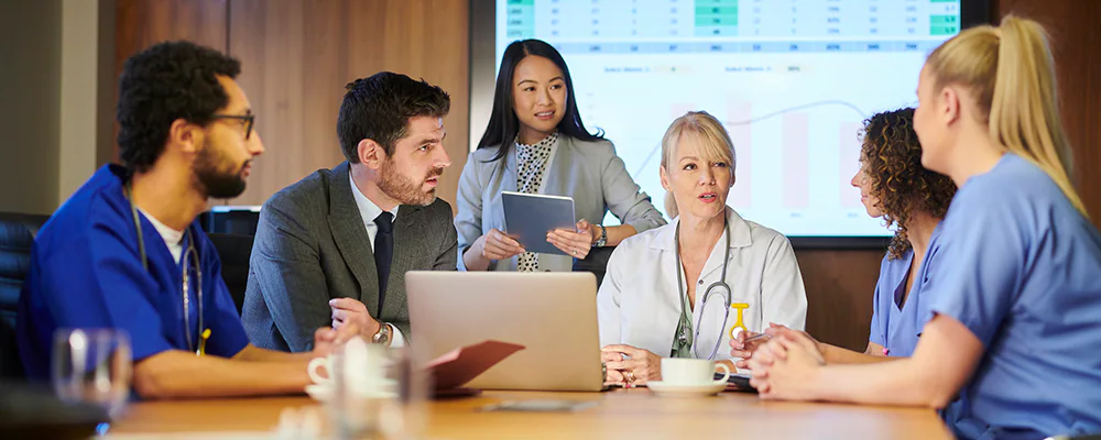 Team meeting between various staff members including physicians, patient care team, and hospital administration showing graphs with data on a screen in background.