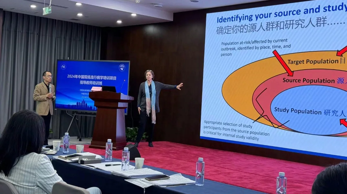 Two presenters standing at the front of a conference room giving a lecture. A woman is pointing to a slide projected onto a screen. The slide on the screen is titled "Identifying your source and study population".