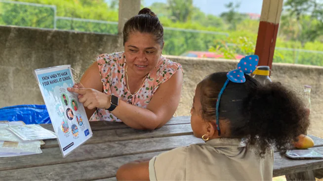 An adult sits across from a young student and points to a poster that reads "Know When to Wash Your Hands at School."