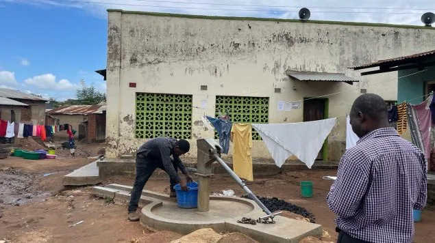 Two individuals stand outside. One gathers water in a bucket while the other looks down.