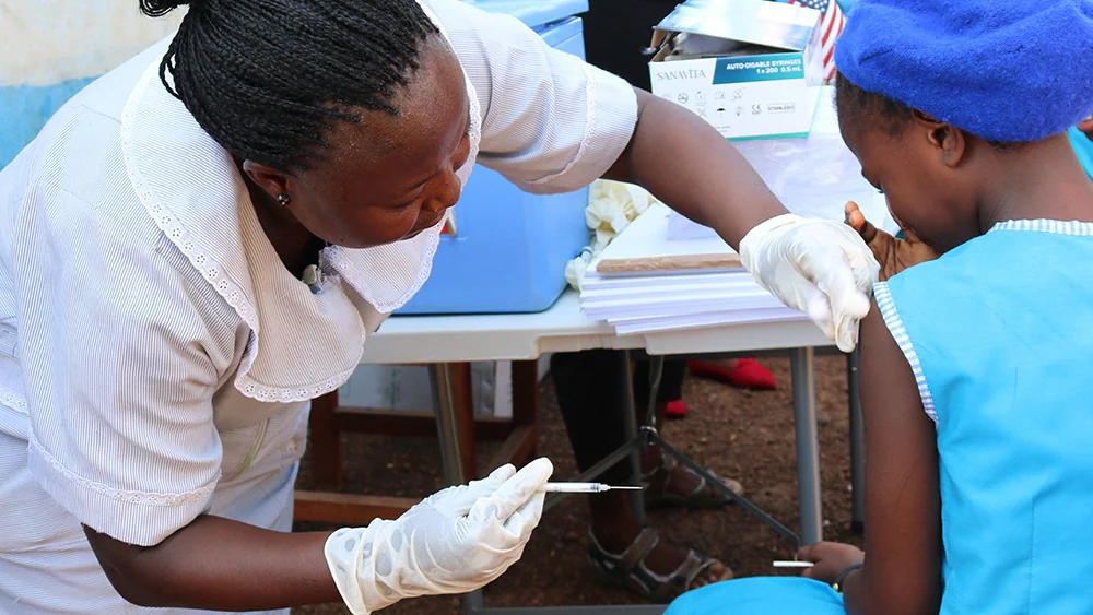 — title missing — A health worker in a white outfit administers a vaccine to a young student who is wearing a blue school uniform.