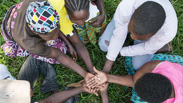 A Community group boosts access to HIV treatment, sitting in a circle, hand stacked, middle hand on top holding a pill