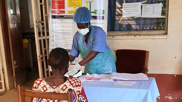 A health worker in personal protective equipment stands behind a desk as she screens a patient in front of her.