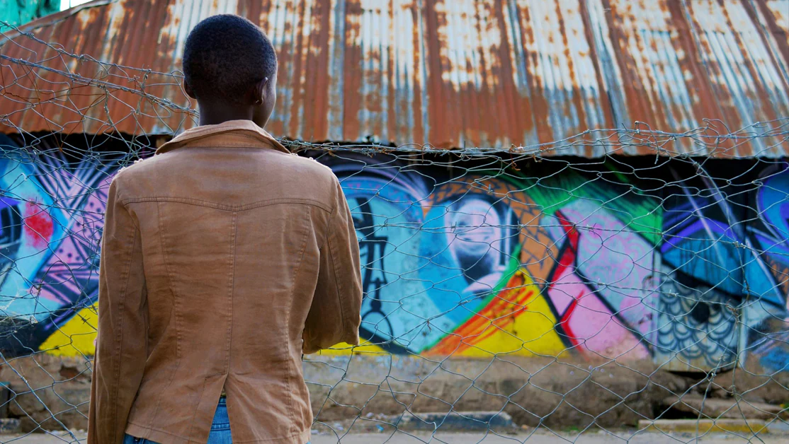 A woman in a brown jacket stands in front of a building with graffiti.