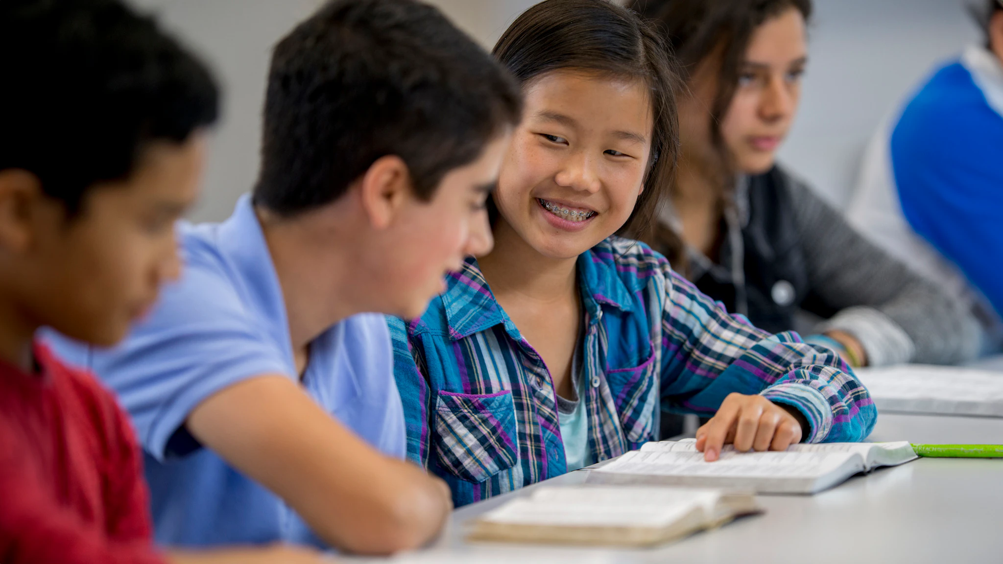 Four students at a table, open books in front of them. Two students in the middle are talking to each other.