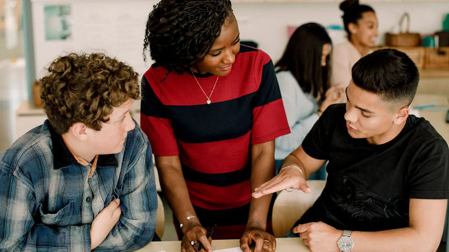 Teacher standing between two seated students, listening to them as they speak.