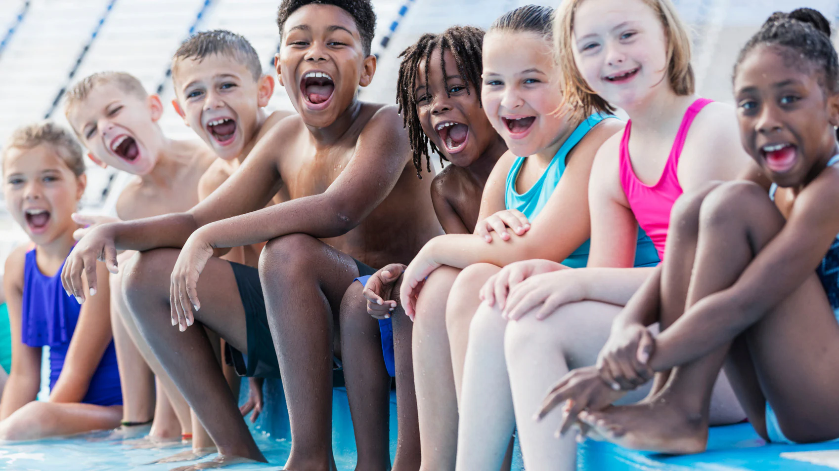 A group of ethnically diverse children sitting in a line at the edge of a pool in their bathing suits laughing.