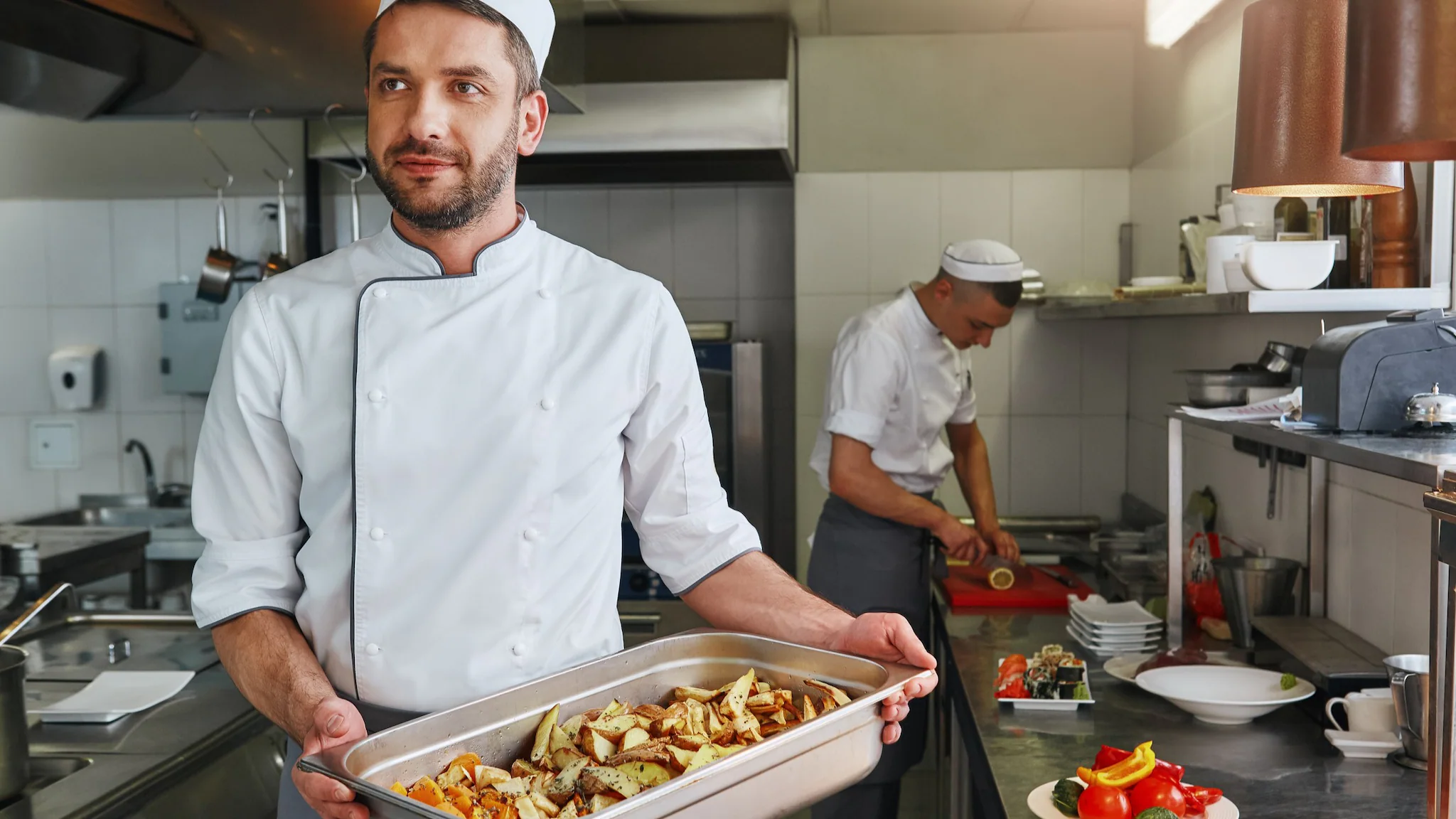 Food workers in a kitchen. One has a large pan of food.