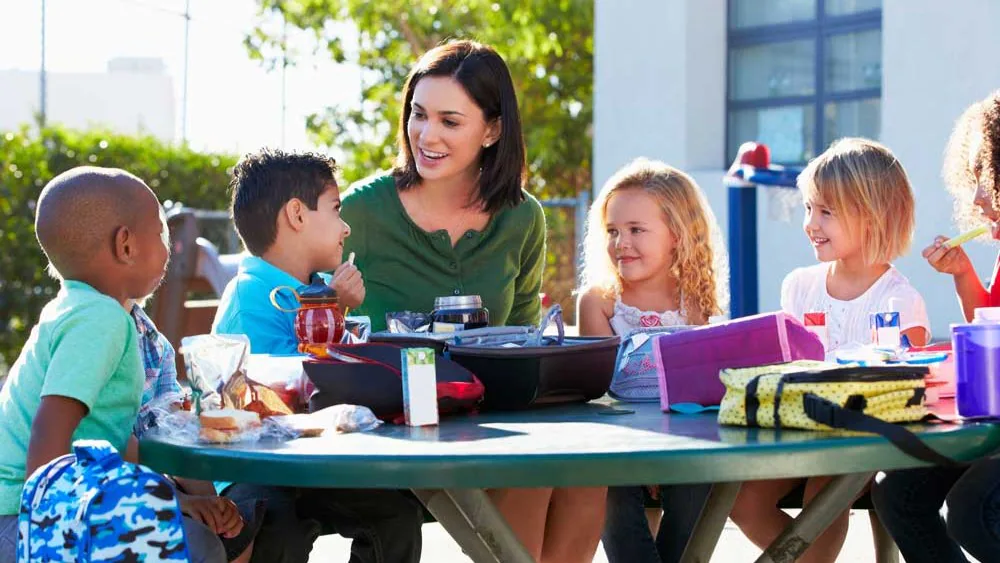 Adult and five children at table eating a meal outside.