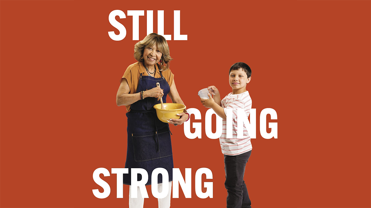 An older woman, wearing an apron, is holding a bowl and using a wooden spoon to stir contents of the bowl. Her adolescent grandson is holding a measuring cup and is about to pour its contents into the bowl.