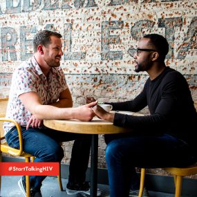 Image displays two men sitting inside holding hands while gazing at each other.