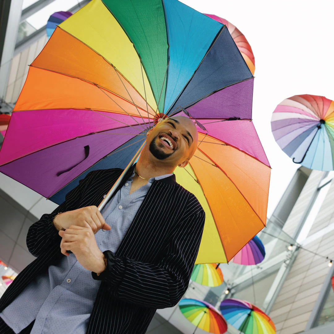 An image of a man smiling outdoors with rainbow umbrellas in the background.