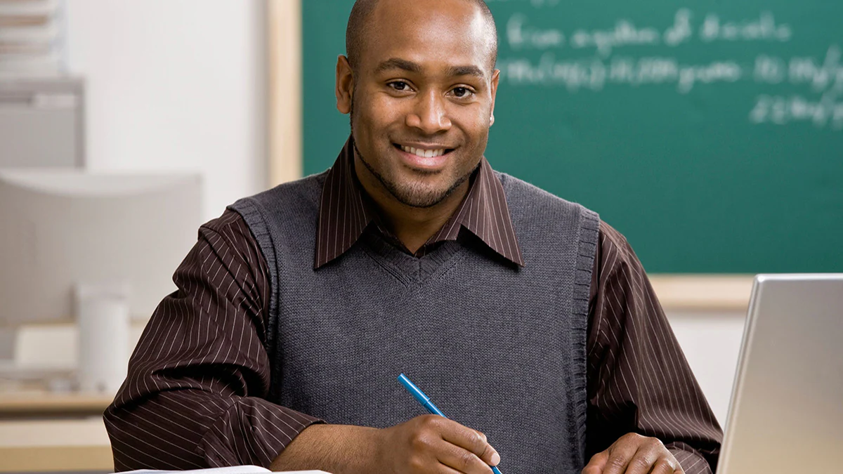 A teacher sits on a desk, holding a notebook in his hands.