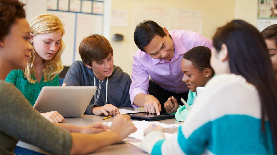 Students sitting around a table with two teachers reviewing schoolwork.